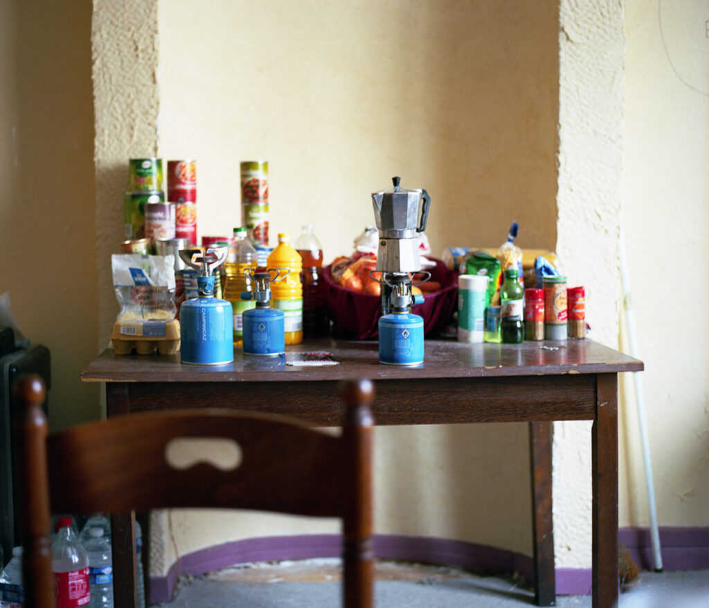 Une table en bois remplie de provisions dans un squat (cafetière, rechaud, conserves...). Photographie issue du projet ZONE 54