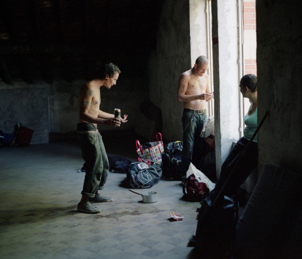 Deux hommes et une femme près d'une grande fenêtre dans un squat insalubre. A leurs pieds quelques affaires. Photographie issue du projet ZONE 54