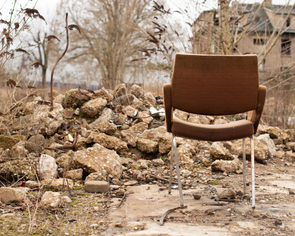 Chaise isolée au milieu de gravats et de végétation, devant un bâtiment industriel en ruine, scène d’abandon sur une friche industrielle en France, trace de présence humaine disparue.