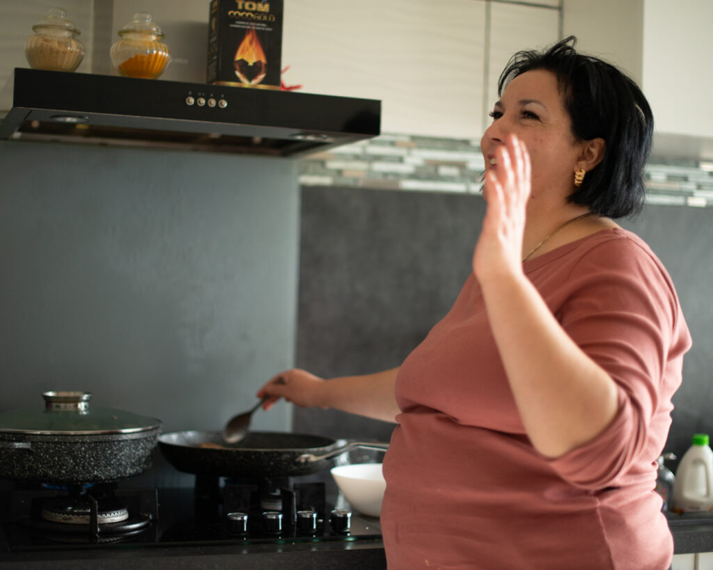 Femme aux cheveux noirs au carré et haut rose, riant en cuisinant à la poêle sur une gazinière, une spatule à la main. Cuisine équipée d'une hotte noire et d'étagères garnies de bocaux d'épices. Convivialité et cuisine maison, série Persévérantes.