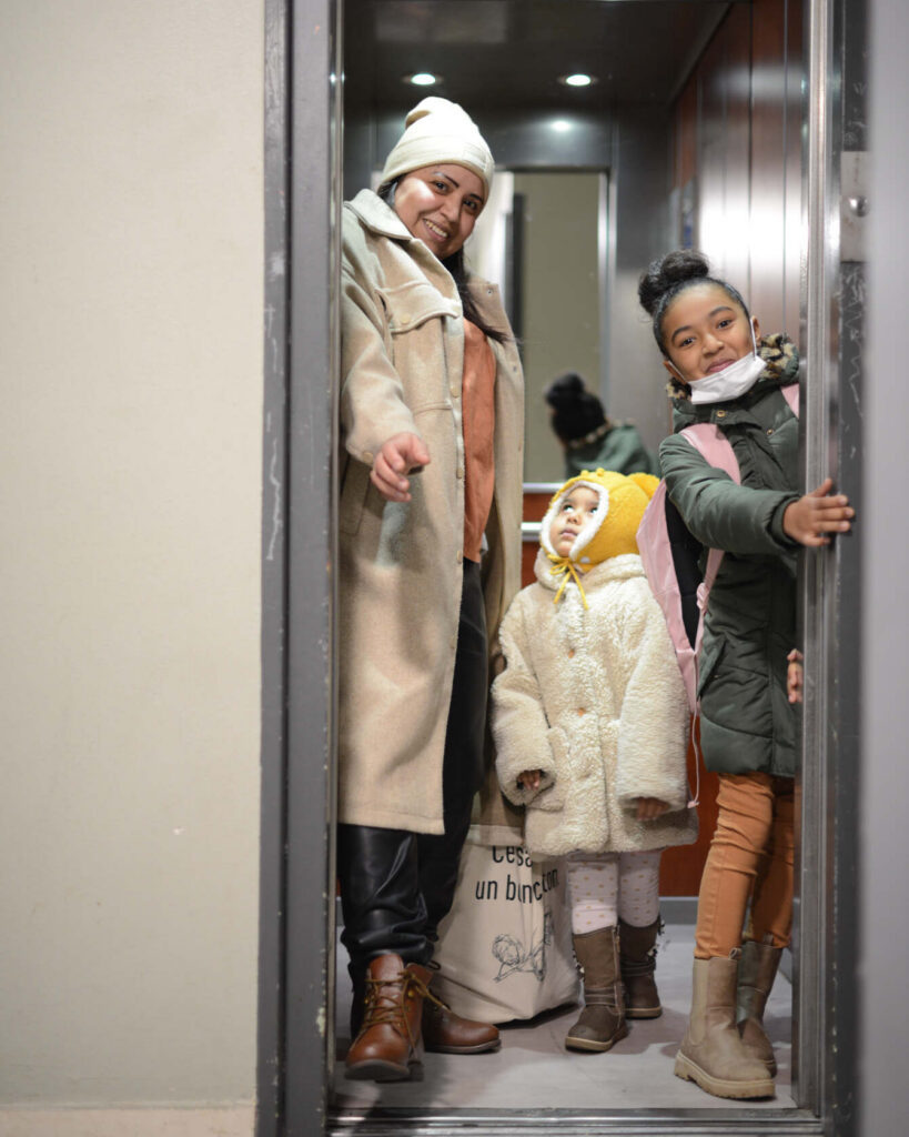 Femme souriante en bonnet et long manteau beige, tenant un sac en toile, accompagnée de deux fillettes emmitouflées dans des manteaux d'hiver, dont une au bonnet jaune et l'autre au cartable rose. Toutes trois posent dans un ascenseur. Sortie en famille, série Persévérantes.