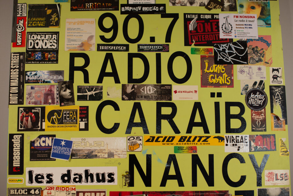 Mur jaune recouvert d’affiches et de stickers avec l’inscription « Radio Caraïb Nancy 90.7 », trace d’une vie culturelle et musicale locale au Haut-du-Lièvre.