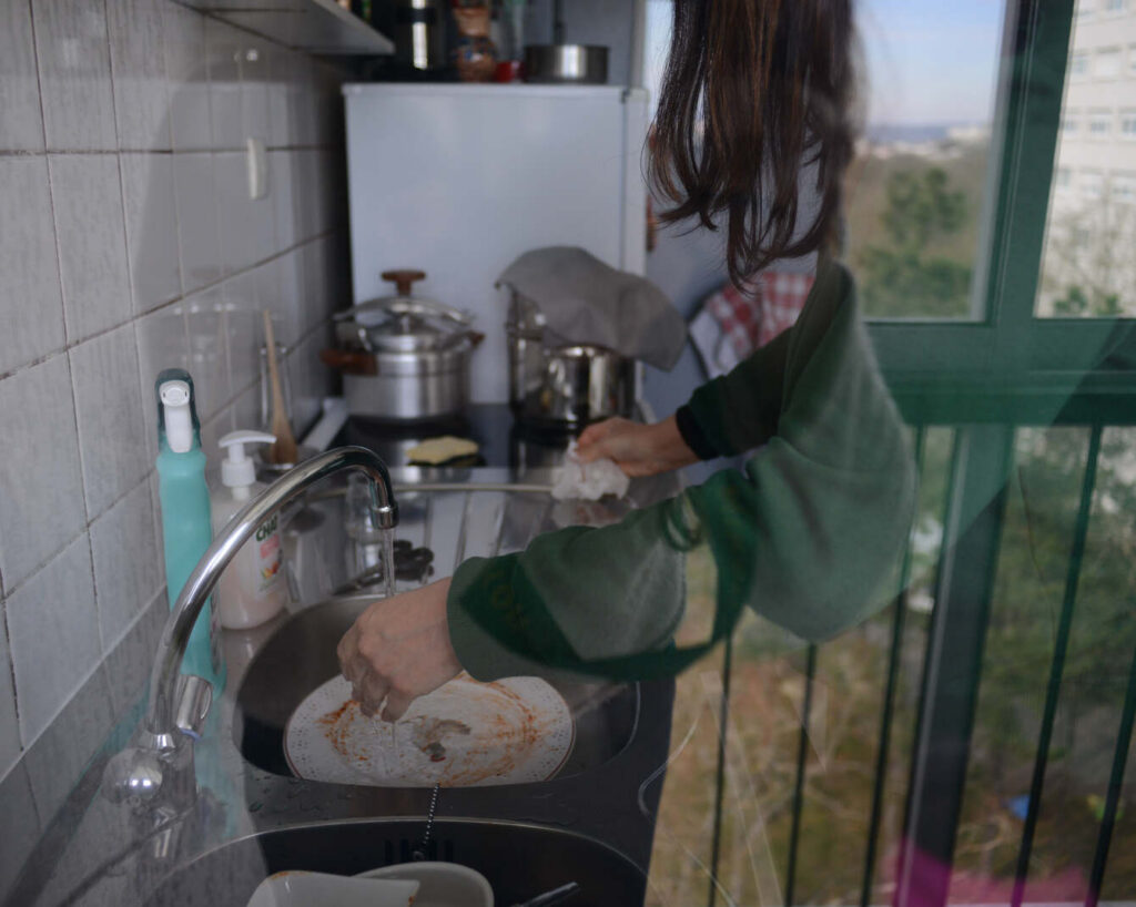 Femme de dos aux longs cheveux bruns et au sweat vert, lavant une assiette tachée de sauce sous l'eau courante d'un évier. Cuisine équipée de cocottes en inox, vue sur les immeubles et la verdure du plateau de Haye par la fenêtre à barreaux verts. Travail domestique quotidien, série Persévérantes
