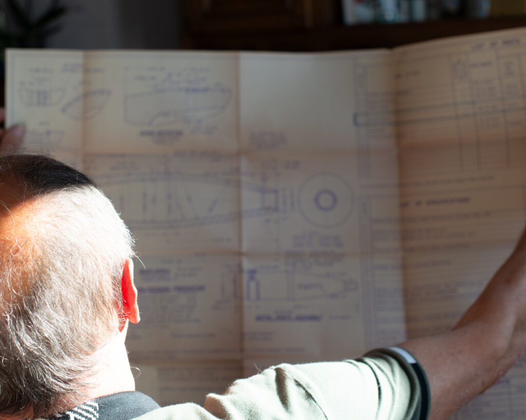 Vu de dos, un homme aux cheveux gris déployant à bout de bras un grand plan technique annoté de schémas et de tableaux, éclairé par une lumière rasante. Mémoire industrielle et transmission par un·e ancien·ne ouvrier·e, série Trifolium Hybridum, Thionville.