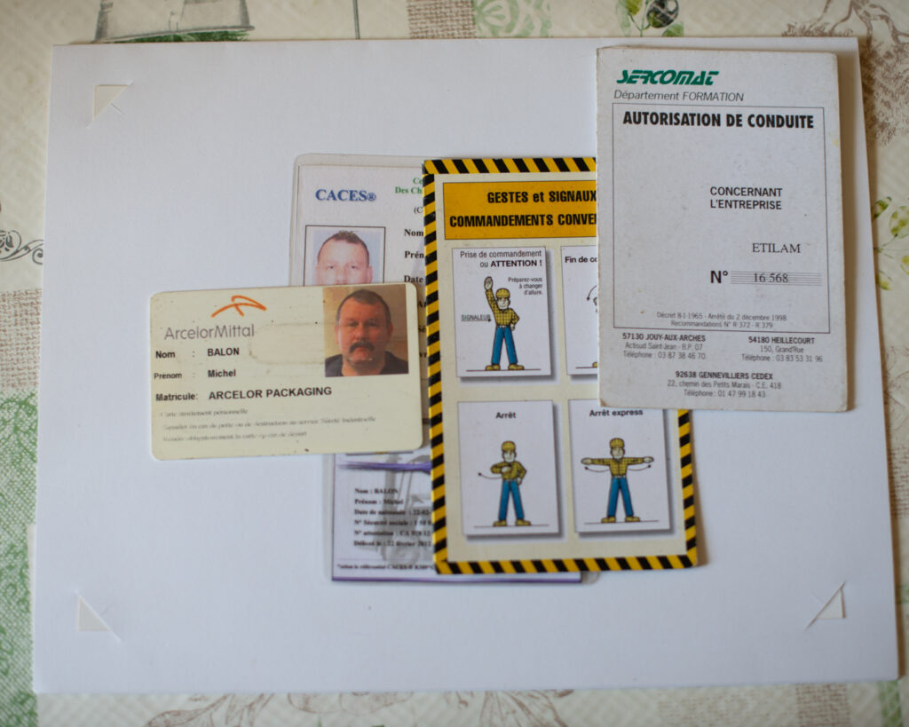 Documents professionnels disposés sur une nappe : badge ArcelorMittal au nom de Michel Balon, carte CACES, fiche illustrée de gestes et signaux conventionnels et autorisation de conduite Sercomat. Archives personnelles d'un·e ancien·ne ouvrier·e des Laminoirs, série Trifolium Hybridum, Thionville.