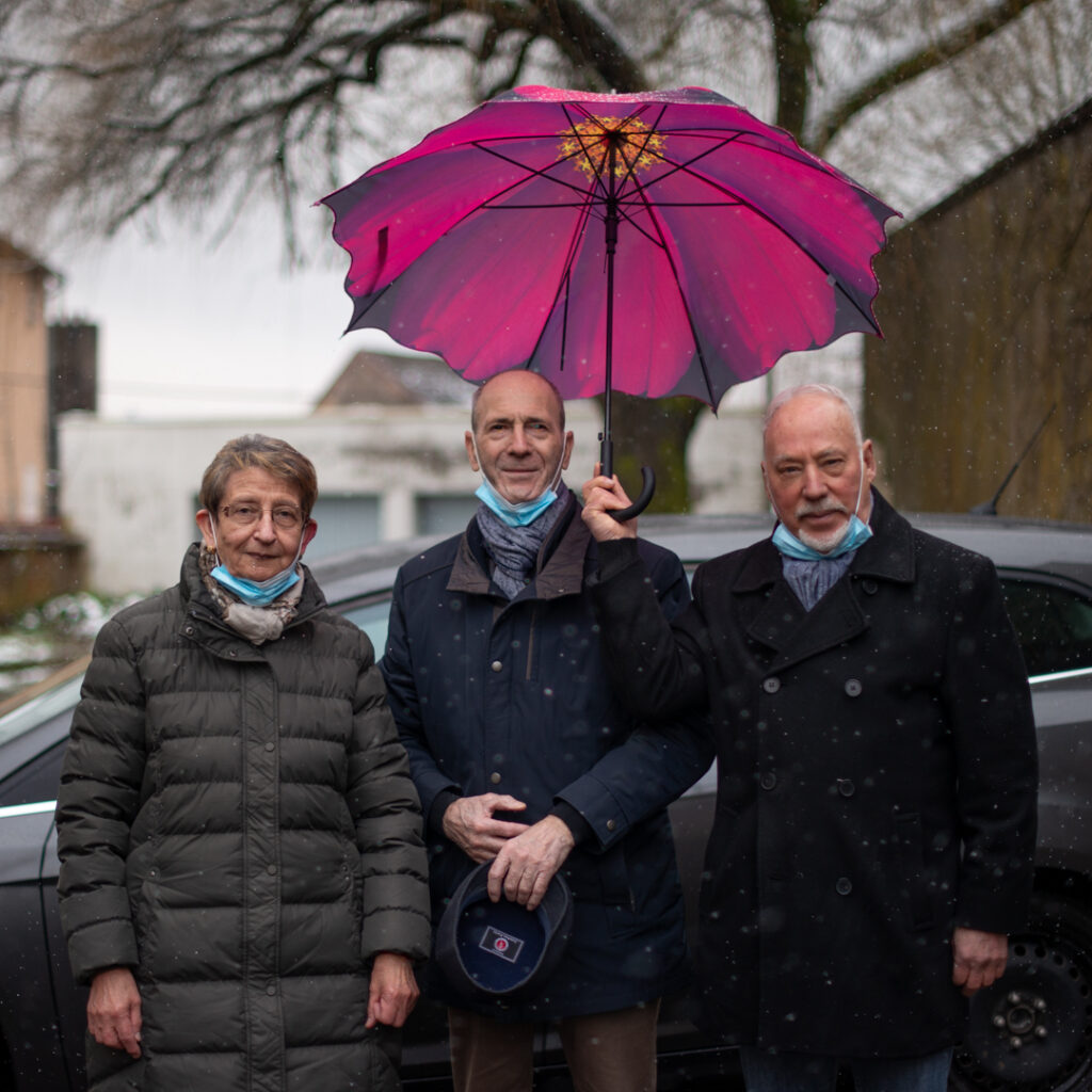 Trois personnes posant sous un parapluie rose dans la rue, portrait collectif d’habitant·e·s, moment de sociabilité et de quotidien partagé.