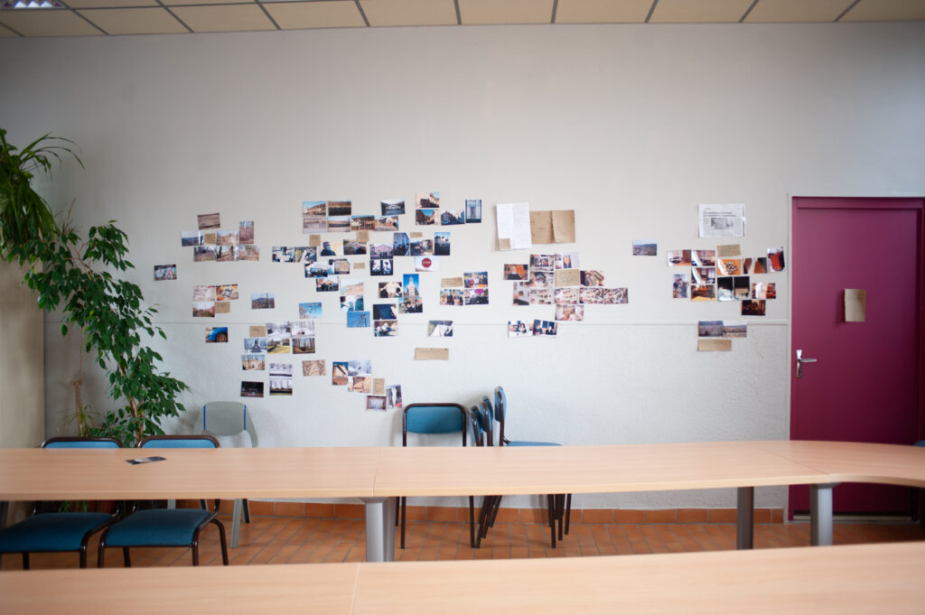 Salle avec tables et mur couvert de photographies et documents, espace de travail collectif autour de la mémoire, archives et récits d’habitant·e·s de Longwy.