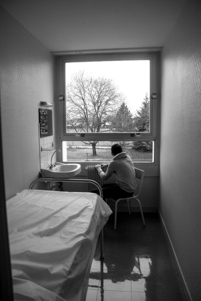 Chambre d’hôpital reconstituée : lit couvert, fenêtre sur la cour, un patient assise près de la fenêtre