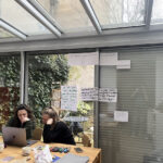 Deux femmes devant un ordinateur. Sur le mur derrière sont accrochés les graffitis réalisés par d'autres. Des marqueurs, feutres, stylo, sont disposés sur la table