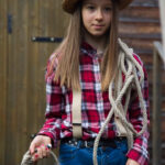 Adolescente en chapeau de paille, chemise à carreaux et gilet, tenant une corde en forme de lasso, et posant dans la cour de la ferme. Mise en scène qui rappelle la paysannerie nord américaine