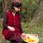 Adolescente en robe rouge et voile à fleurs, agenouillée au bord d'un chemin, cueillant des pommes dans un petit panier en osier. Mise en scène qui rappelle l'un de se souvenirs en algérie