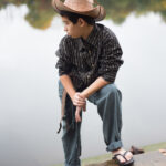 adolescend portant un chapeau et des vêtements d’inspiration paysanne, debout près d’un point d’eau (lac ou étang), posture concentrée, lumière douce et arrière-plan végétal flou. Mise en scène qui rappelle les rizières.