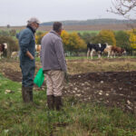 Deux personnes en bottes, près d’un enclos clôturé, discutant au bord d’un terrain labouré ; vaches en arrière-plan dans le pré