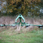 Tracteur et outil agricole vert sur une prairie couverte d’herbes hautes, au milieu d’une végétation de bordure et d’arbres ; activité de préparation et d’entretien de parcelle en milieu rural.