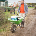 Homme en tenue de travail poussant une brouette chargée de matériel dans une allée de ferme, avec en arrière-plan des installations agricoles et une végétation de saison.