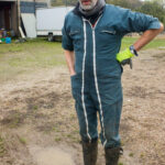 Agriculteur en combinaison de travail, gants en latex et casquette, au milieu de la cour de la ferme, devant des bâtiments agricoles et des équipements ; scène de vie à la ferme biologique et de quotidien paysan.