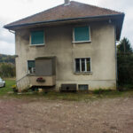 Maison de ferme sobre et enduite, façades grises, fenêtres et portes vertes, sol en terre près de la bâtisse et paysage rural autour.