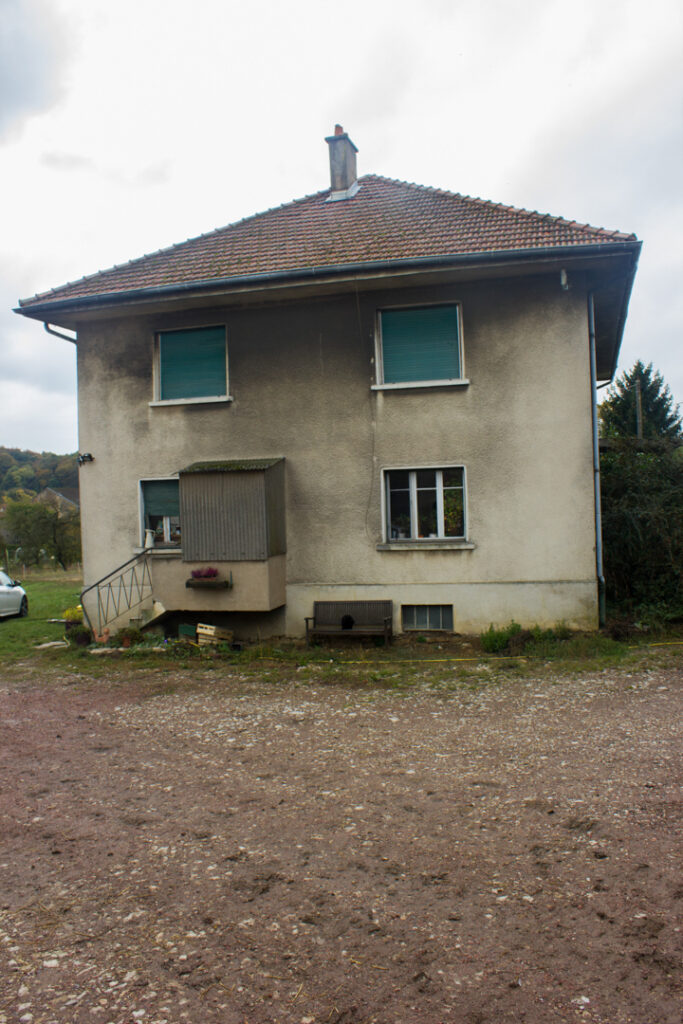 Maison de ferme sobre et enduite, façades grises, fenêtres et portes vertes, sol en terre près de la bâtisse et paysage rural autour.
